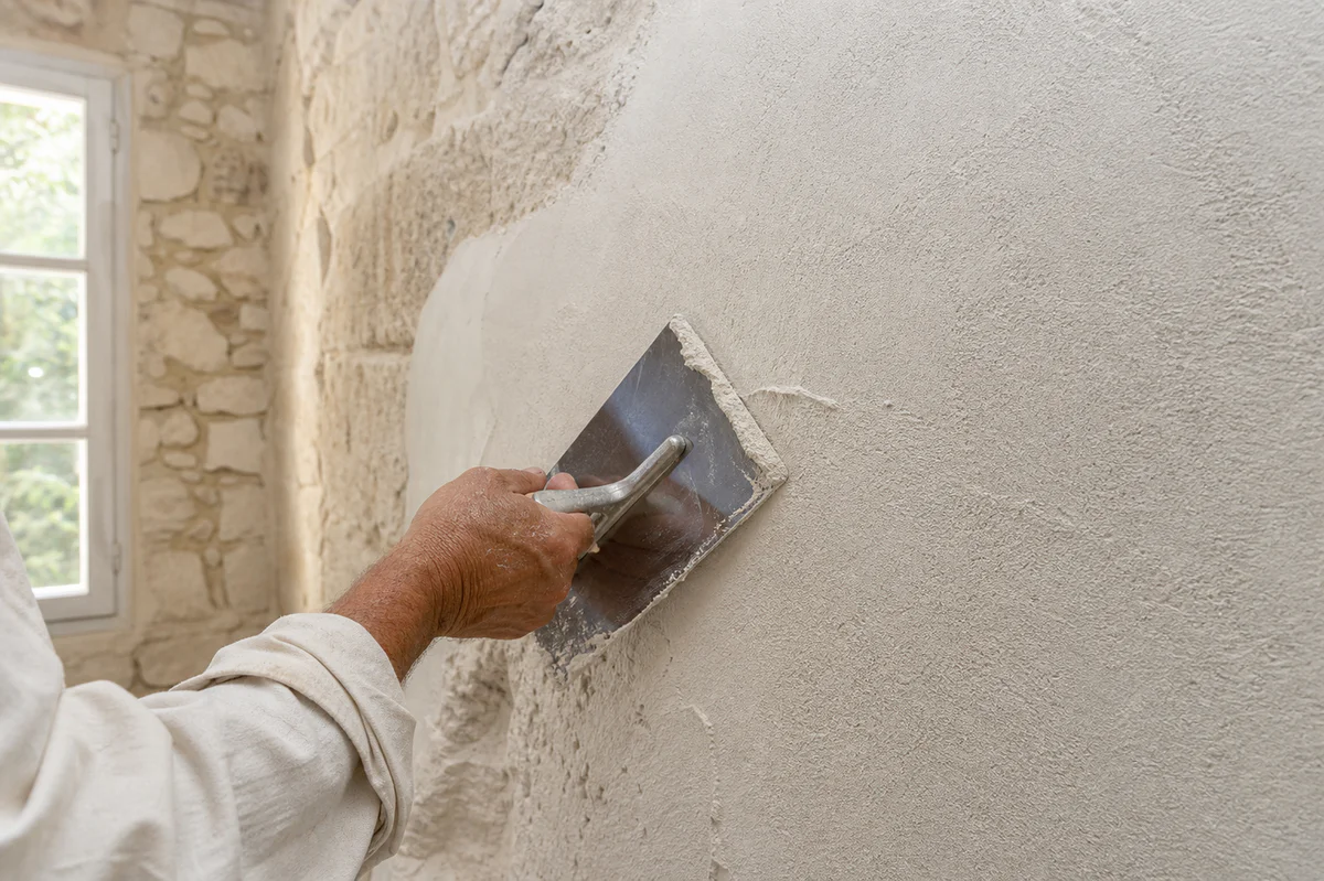 Artisan applying lime plaster to an old interior stone wall in a warm traditional Provence renovation setting.