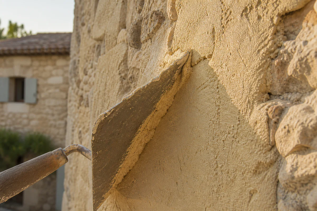 Artisan appliquant un enduit à la chaux naturelle premium sur un mur en pierre calcaire provençal, texture fine et lumière ch